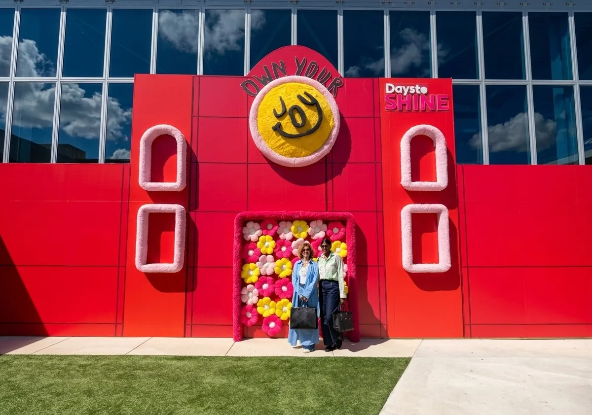 Vivianka Lavin and Yaima Ortiz posing in front of a red wall with flowers and an Own Your Joy sign