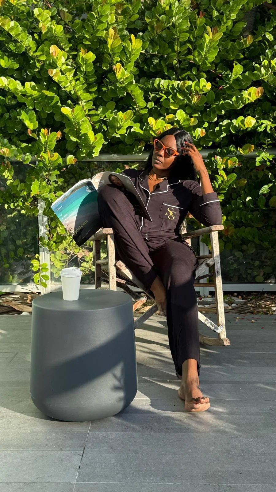 A woman relaxing outdoors in the sun wearing the black Spry pyjama set with gold monogram embroidery