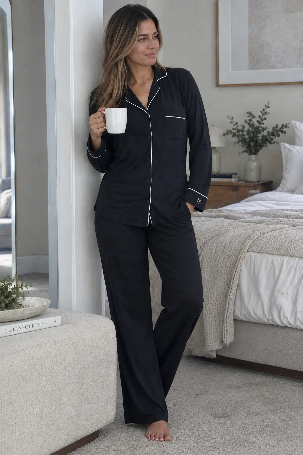 A woman in the black Spry pyjama set with ivory piping, coffee in hand, in a calm bedroom