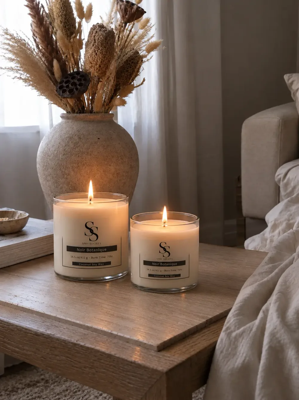 Two lit Spry Society Noir Botanique candles on a wooden side table beside dried flowers